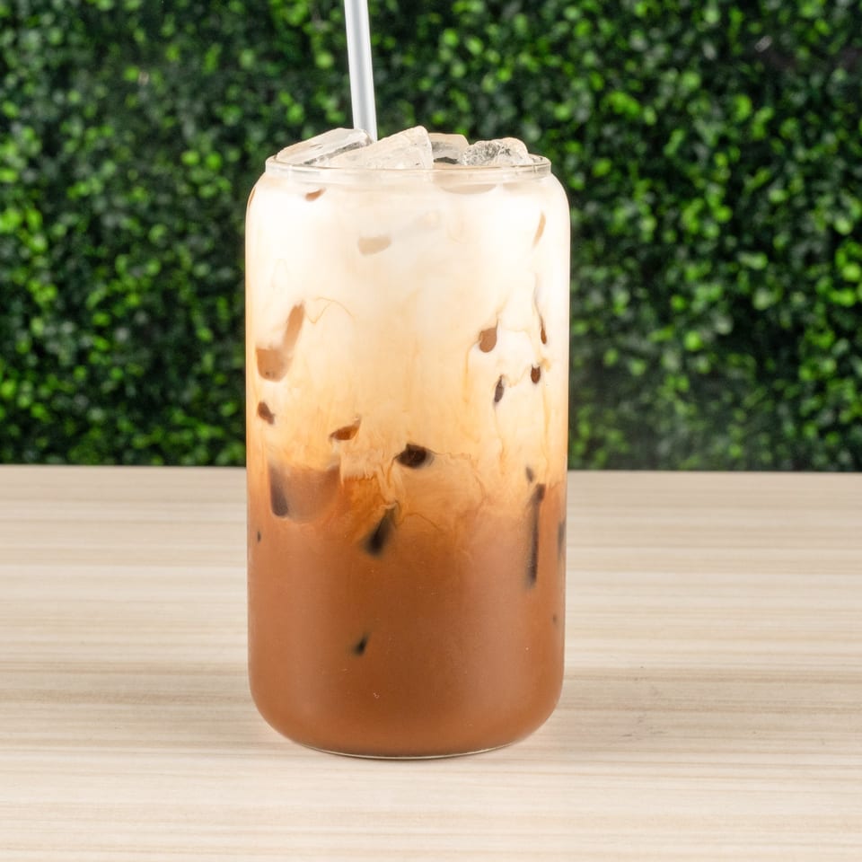 Iced layered brown and white drink with a straw, on wood against green foliage.