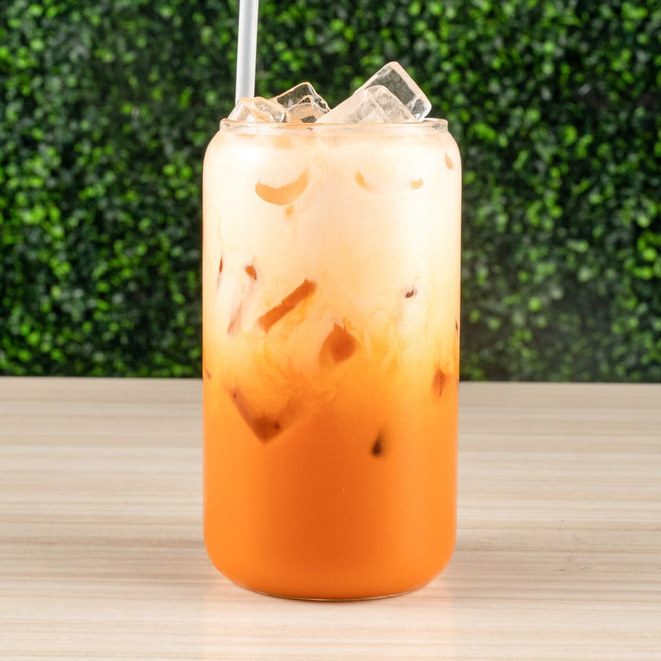 A tall glass of layered Thai iced tea with ice and a straw on a light wood surface.