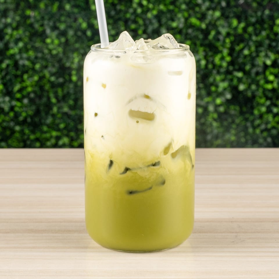 Iced green tea latte with a creamy white top layer and a straw on a wooden table.
